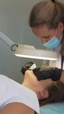 Doktor, kadının yüzündeki sivilceleri çıkartıyor. Siyah eldivenli eller steril süngerleri tutar ve açık tenli sivilceleri temizler. Lamba ve büyüteç, yüzü aydınlatır. Cilt temizleme işlemi