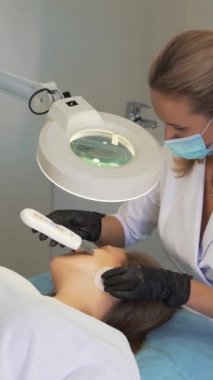 Kozmetik uzmanı deriyi ultrasonik temizlik cihazıyla temizliyor. Eldivenli doktorlar müşterinin yüzünü metal plakayla kaplıyor. Yenilenme prosedürü, sivilce ve sivilce tedavisi