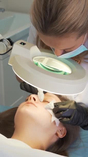 Doctor squeezes out pimples on face of woman. Hands in black gloves hold sterile sponges and remove dirt and pimples from fair skin. Lamp and magnifying glass illuminate face. Skin cleansing procedure — 图库视频影像