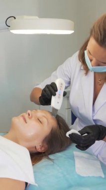 Kozmetik uzmanı deriyi ultrasonik temizlik cihazıyla temizliyor. Eldivenli doktorlar müşterinin yüzünü metal plakayla kaplıyor. Yenilenme prosedürü, sivilce ve sivilce tedavisi