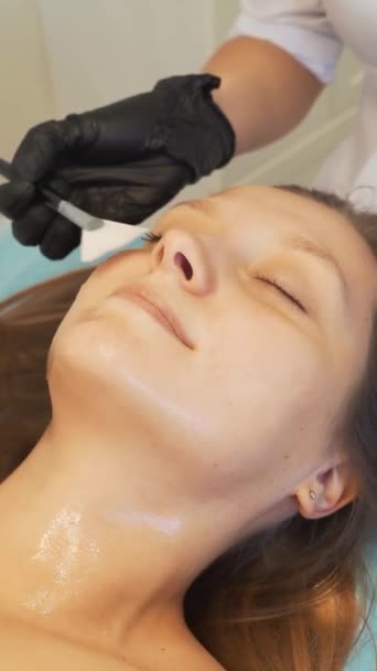 Kosmetolog nakłada maskę na skórę kobiecej twarzy w celach terapeutycznych. Pacjent leży na kanapie, a grubą przezroczystą substancję nakłada się na twarz pędzlem. Odmładzanie, leczenie trądziku — Wideo stockowe