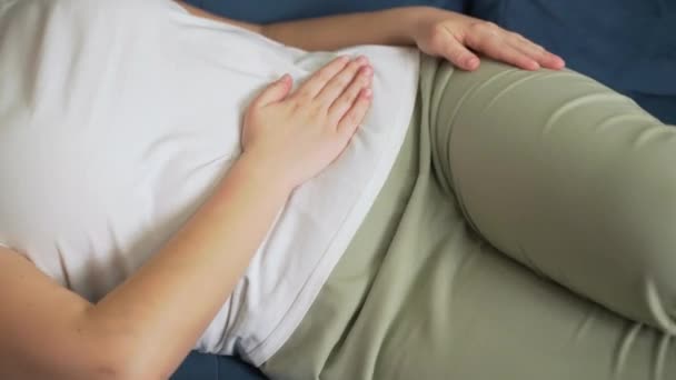 Жінка в білій футболці лежить на синьому дивані і тримає живіт рукою. Інсульт шлунку рукою, щоб він не постраждав. Менструація, вагітність, діарея, задишка. Біль на обличчі — стокове відео