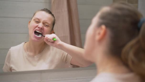 La mujer de camiseta beige se cepilla activamente los dientes con un cepillo de dientes verde en el baño de su casa. Limpia los dientes de la suciedad y los alimentos para la salud dental. Dientes sanos, sonrisa hermosa, rutina diaria — Vídeos de Stock