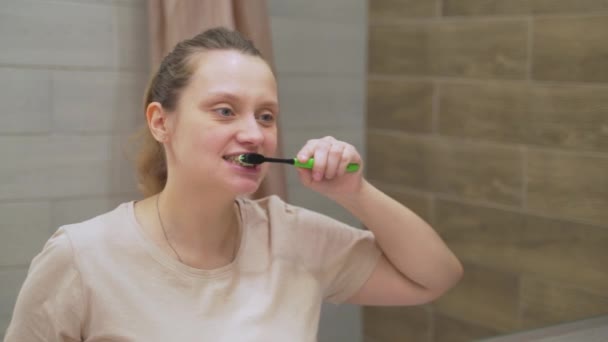 Bej tişörtlü kadın evdeki banyoda dişlerini yeşil diş fırçasıyla fırçalıyor. Topraktan diş temizliği ve diş sağlığı için yiyecek topluyor. Sağlıklı dişler, güzel gülücük, günlük rutin — Stok video