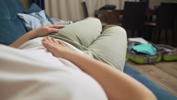 Kvinna i vit T-shirt ligger på blå soffa och håller magen med handen. Slag magen med handen så att det inte gör ont. Menstruation, graviditet, diarré, frosseri. Smärta i ansiktet — Stockvideo