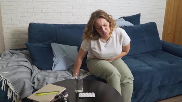Młoda kobieta leży na niebieskiej kanapie, masuje żołądek z powodu silnego bólu i pije wodę. Menstruacja, ciąża, obżarstwo. Na stole jest szklanka wody i pigułki przeciwbólowe.. — Wideo stockowe
