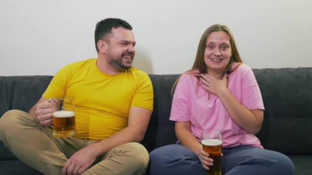 Pareja joven está sentada en un sofá gris, viendo la televisión y bebiendo cerveza de vasos de vidrio transparentes. Buen humor. Están sorprendidos, riendo y sonriendo. Relajarse, enamorarse, ver la televisión — Vídeos de Stock