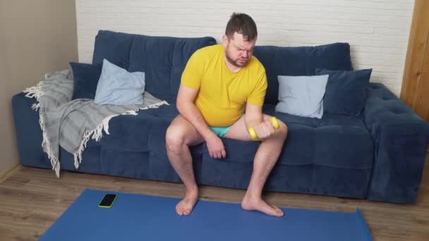 O homem gordo engraçado envolve-se ativamente na aptidão com pequenos halteres amarelos. Atleta realiza ativamente exercício para bíceps e sorrisos. Alegria, auto-ironia. Perda de peso, esportes, auto-isolamento — Vídeo de Stock