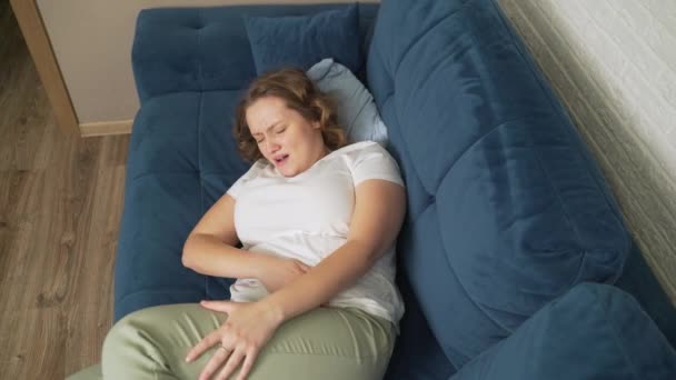 Mujer en camiseta blanca está acostada en un sofá azul y sosteniendo el estómago con la mano. Ella acaricia el estómago con la mano para que no duela. Menstruación, embarazo, diarrea, gula. Hay dolor en la cara. — Vídeos de Stock