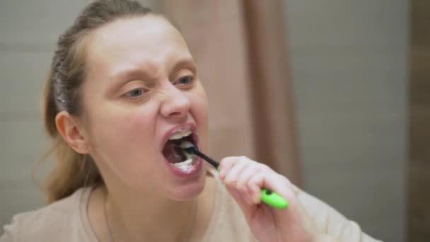 La mujer de camiseta beige se cepilla activamente los dientes con un cepillo de dientes verde en el baño de su casa. Limpia los dientes de la suciedad y los alimentos para la salud dental. Dientes sanos, sonrisa hermosa, rutina diaria — Vídeo de stock