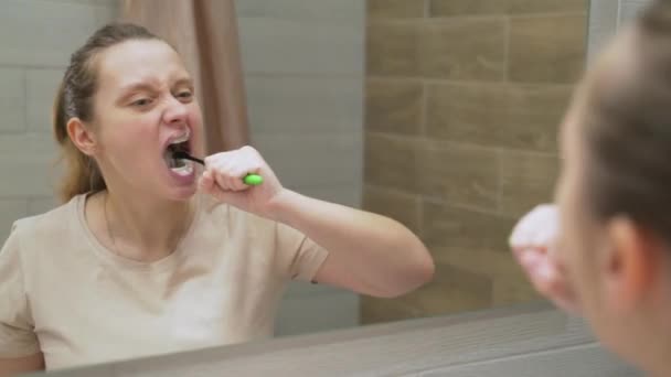 Женщина в бежевой футболке активно чистит зубы зеленой зубной щеткой в ванной комнате дома. Она очищает зубы от грязи и пищи для стоматологического здоровья. Здоровые зубы, красивая улыбка, ежедневная рутина — стоковое видео