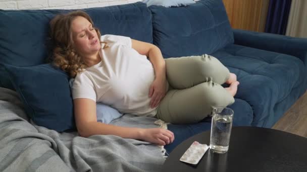 Frau liegt auf Sofa und massiert Bauch mit der Hand vor starken Schmerzen und Krämpfen. Auf einem dunklen Holztisch liegen Wassergläser und Schmerztabletten. Menstruation, Schwangerschaft, Völlerei, Schmerzen, Krämpfe — Stockvideo