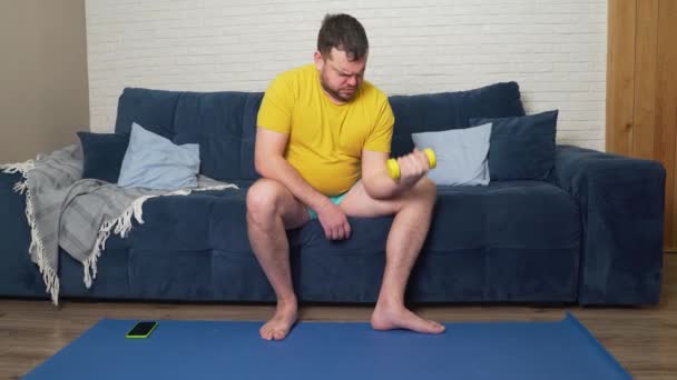 Homme gras drôle est activement engagé dans la remise en forme avec de petits haltères jaunes. Athlète effectue activement des exercices pour les biceps et les sourires. Joyeux, ironie de soi. Perte de poids, sport, auto-isolement — Video