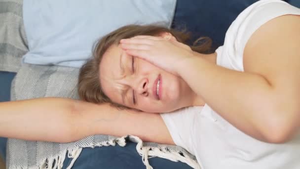 Mladá žena leží na posteli se zavřenýma očima a masíruje spánky hlavy kvůli migréně. Bolí ji hlava. Na její tváři je utrpení a bolest. nachlazení, chřipka, migréna, epidemie, křeče — Stock video