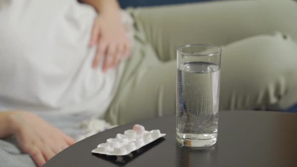 Hay un vaso de agua y pastillas para el dolor en la mesa de madera oscura. La mujer se acuesta en el sofá y masajea el estómago con la mano de dolor severo y calambres. Menstruación, embarazo, glotonería, dolor, calambres — Vídeo de stock