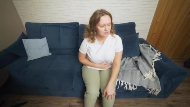 Mulher de t-shirt branca está sentada no sofá azul e segurando o estômago com a mão na dor. Menstruação, gravidez, diarreia, gula. O rosto está a ficar com dores. — Vídeo de Stock