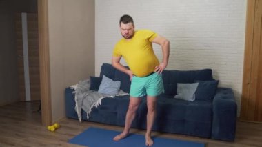 Şişman sakallı adam garip bir şekilde ve özenle evde kalçasını çeviriyor. Evde kilo vermek ve ısınmak için fitness antrenmanı yapıyor. Sağlıklı yaşam tarzı, kilo kaybı, izolasyon