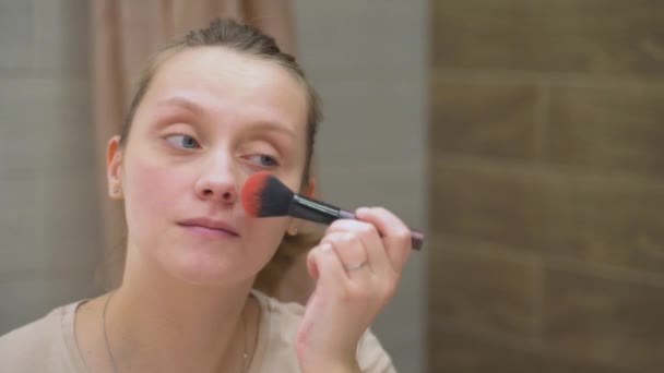 Femme aux yeux bleus pulvérise la peau sur le visage avec une brosse moelleuse, debout par un grand miroir dans la salle de bain. Ses cheveux ondulés sont attachés dans une queue de cheval. Matin, maquillage, réveil — Video