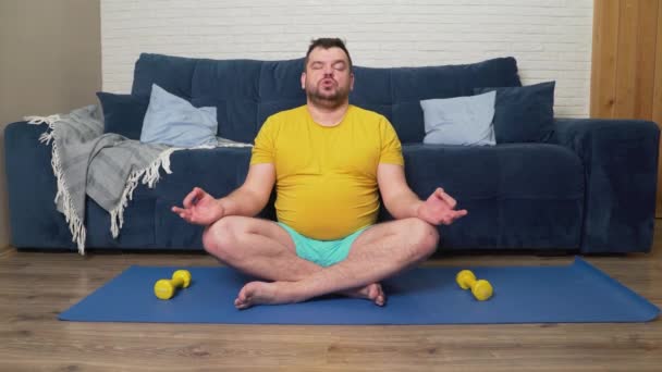 El hombre de camiseta amarilla está sentado en la alfombra en la posición de loto y meditando. Hace yoga. Inhala y exhala lenta y uniformemente. Recuperación, relajación, conciencia, meditación, yoga — Vídeos de Stock