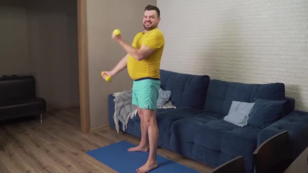 Hombre gordo divertido se dedica activamente a la aptitud con pequeñas mancuernas amarillas. Atleta realiza activamente ejercicio para bíceps y sonrisas. Alegría, autoironía. Pérdida de peso, deportes, autoaislamiento — Vídeo de stock