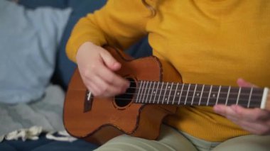 Ukulele 'de şarkı performansı. Bir el gitar tellerini ritme göre nazikçe çırpıyor, diğer elin parmakları enstrümanın boynuna klemp takıyor. Müzik okulu, online eğitim.