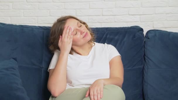 Egy fiatal nő ül az ágyon, migrénje van. Súlyos görcsök miatt masszírozza a feje halántékát. Szenvedve az arcán, mélyen lélegzik. Hideg, influenza, migrén, járvány, görcsök — Stock videók