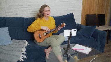 Kadın çevrimiçi bir ders veriyor ve öğrencilere ukulele çalmayı öğretiyor. Gitarı elinde tutuyor ve insanlara canlı video bağlantısıyla anlatıyor. Bloglama, tecrit, çevrimiçi eğitim