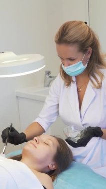 Kozmetik uzmanı kadın derisine tedavi amaçlı maske takıyor. Hasta kanepede yatıyor ve kalın şeffaf madde fırçayla yüzüne uygulanıyor. Gençleştirme, sivilce tedavisi