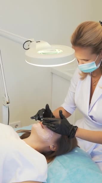 Cosmetologist applies white mask to fair skin of womans face in circular movements before procedures. Cleansing skin of impurities. Doctor has gloves on his hands. Skin cleansing, acne treatment — Stockvideo