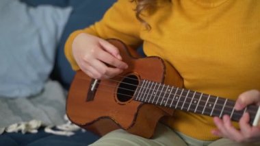 Ukulele 'de şarkı performansı. Bir el gitar tellerini ritme göre nazikçe çırpıyor, diğer elin parmakları enstrümanın boynuna klemp takıyor. Müzik okulu, online eğitim.