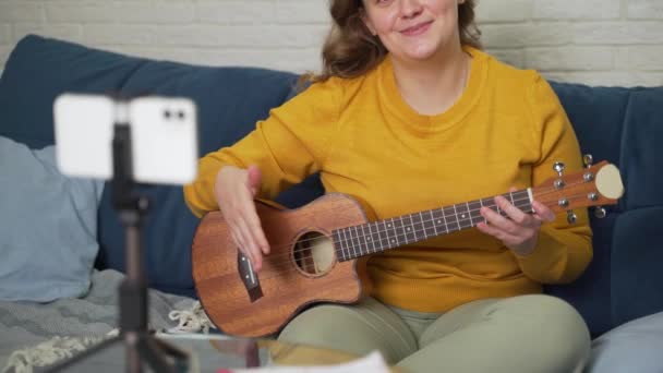 Perempuan melakukan pelajaran online dan mengajar siswa untuk memainkan ukulele. Memegang gitar di tangan dan memberitahu orang-orang tentang hal itu melalui link video langsung. Blogging, isolasi, pelatihan online — Stok Video