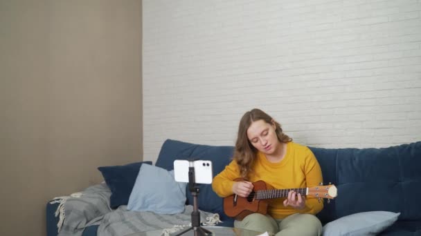 Frau lernt Ukulele spielen, indem sie Video-Tutorial am Telefon anschaut. Online-Gitarrenunterricht. Sie hält die Gitarre in der Hand und versucht, Akkorde zu studieren, während sie aufmerksam auf den Telefonbildschirm blickt. Online-Schulung — Stockvideo