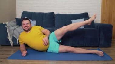 Sarı tişörtlü tombul sakallı adam bacağını seve seve kaldırır ve yan yatar. Eli karnının üstünde. Harika bir ruh hali, evde fitness eğitimi, kilo kaybı, izolasyon