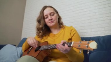 Kadın ukulele 'de melodi çalıyor. El gitar tellerini nazikçe ritme göre çırpar, diğer elin parmakları enstrümanın boynuna klemp takar. Müzik okulu, online eğitim, izolasyon