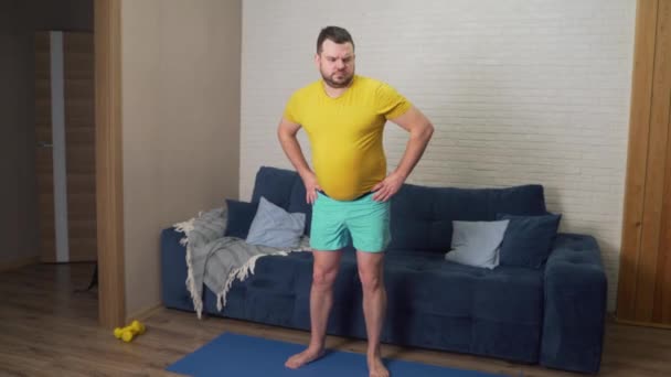 Sarı tişörtlü şişman adam kalçalarını garip ve gayretli bir şekilde hareket ettiriyor. Evde kilo vermek ve ısınmak için fitness antrenmanı yapıyor. Sağlıklı yaşam tarzı, kilo kaybı, izolasyon — Stok video