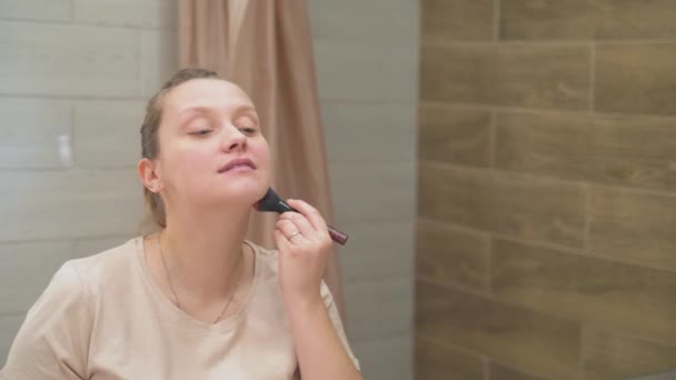 Mujer con ojos azules está empolvando la piel en la cara con un cepillo esponjoso, de pie junto a un gran espejo en el baño. Su cabello ondulado está atado en una cola de caballo. Buenos días, maquillaje, despertar — Vídeo de stock