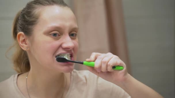 Mulher em camiseta bege ativamente escova os dentes com escova de dentes verde no banheiro em casa. Ela limpa os dentes da sujeira e da comida para a saúde dentária. Dentes saudáveis, belo sorriso, rotina diária — Vídeo de Stock