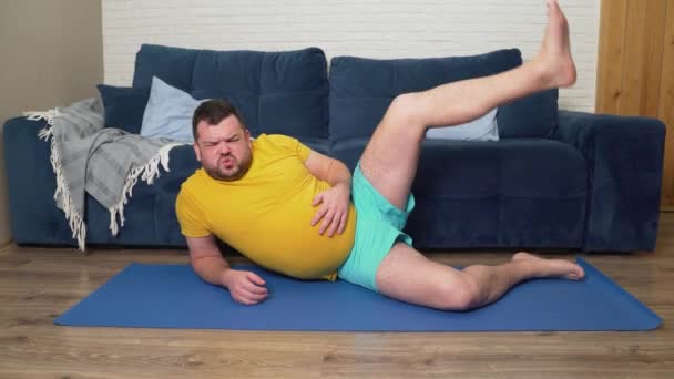 Stout brodaty mężczyzna w żółtej koszulce podnosi nogę, leżąc na boku na macie, jest zgięty na kolanie. Niewłaściwe wykonywanie ćwiczeń sportowych. Ciężko oddycha. Trening w domu, odchudzanie, izolacja — Wideo stockowe