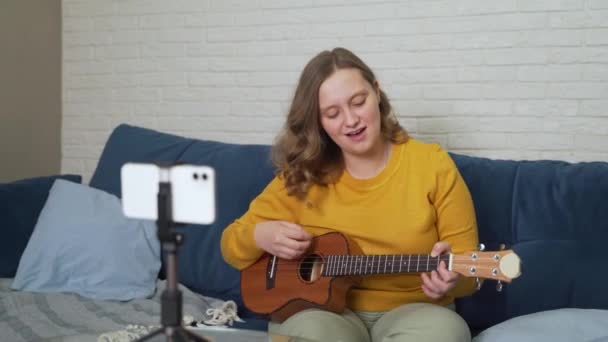 Femme conduit des leçons en ligne, enseignant aux élèves à jouer de l'ukulélé. Joue de la guitare et chante des chansons. Devant elle se trouve le smartphone. Montre comment serrer les cordes et les battre. Blogging, formation en ligne — Video