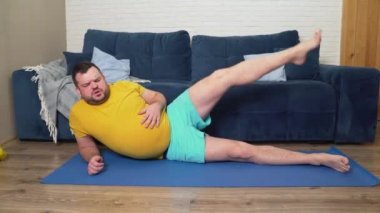 Sarı tişörtlü şişman sakallı adam bacağını kaldırır ve yan yatar. Eli karnının üstünde. Zor nefes alıyor ve etrafa bakıyor. Evde spor eğitimi, kilo kaybı, izolasyon 