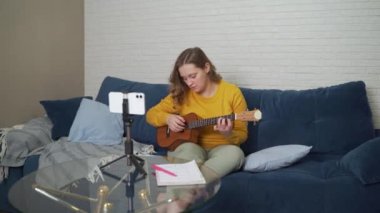 Kadın ukulele çalmayı telefondan video dersi izleyerek öğreniyor. Online gitar dersi. O gitar tutuyor ve müzik akorları üzerinde çalışıyor, dikkatle telefon ekranına bakıyor. Çevrim içi eğitim