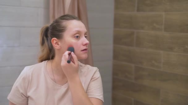 Frau mit blauen Augen pudert die Gesichtshaut mit flauschiger Bürste, steht im Badezimmer vor einem großen Spiegel. Ihr welliges Haar ist zu einem Pferdeschwanz zusammengebunden. Morgen, Make-up, Aufwachen — Stockvideo