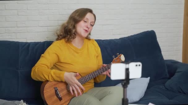 Kadın çevrimiçi bir ders veriyor ve öğrencilere ukulele çalmayı öğretiyor. Gitarı elinde tutuyor ve insanlara canlı video bağlantısıyla anlatıyor. Bloglama, tecrit, çevrimiçi eğitim — Stok video
