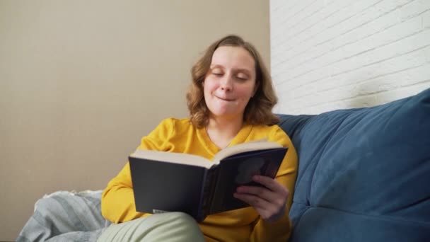 Femme lit avec enthousiasme livre de papier relié assis sur le canapé. Elle feuillette les pages. Elle soupire et hoche la tête du roman qu'elle a lu, sourit. Lecture fascinante, loisirs — Video