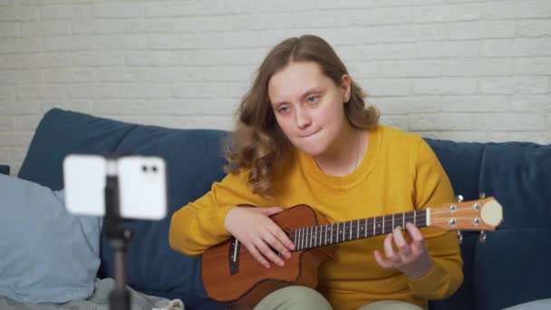 Mujer aprende a jugar el ukelele viendo video tutorial en el teléfono. Clase de guitarra en línea. Ella está sosteniendo la guitarra y tratando de estudiar acordes musicales, mirando atentamente a la pantalla del teléfono. Formación en línea — Vídeo de stock