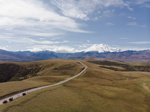 Vista aérea de la increíble carretera con vista a la cresta del Cáucaso y el Monte Elbrus. La autopista atraviesa el prado verde. Nubes claras cubren los picos nevados blancos de Elbrus y el cielo azul Fotos de stock