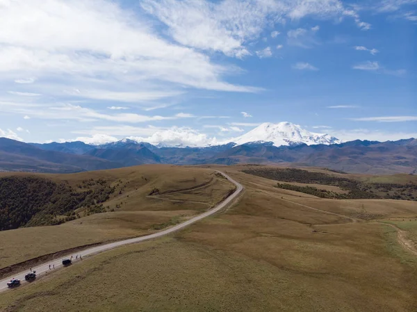 Kafkasya sırtı ve Elbrus Dağı manzaralı harika bir yol manzarası. Otoyol yeşil çayır boyunca uzanıyor. Hafif bulutlar Elbrus 'un beyaz karlı tepelerini ve mavi gökyüzünü kaplıyor.