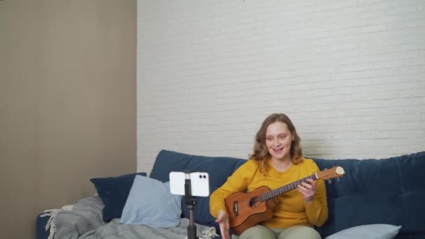 Mujer lleva a cabo una lección en línea y enseña a los estudiantes a tocar el ukelele. Sostiene la guitarra en las manos y le dice a la gente sobre ella a través de enlace de vídeo en vivo. Blogging, aislamiento, formación en línea — Vídeos de Stock
