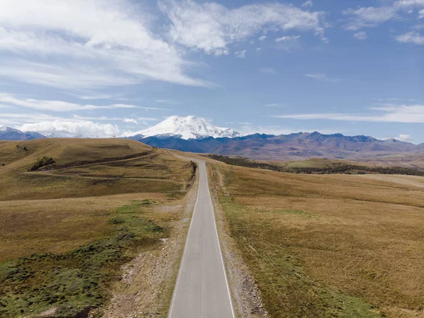 Kafkasya sırtı ve Elbrus Dağı manzaralı harika bir yol manzarası. Otoyol yeşil çayır boyunca uzanıyor. Hafif bulutlar Elbrus 'un beyaz karlı tepelerini ve mavi gökyüzünü kaplıyor.