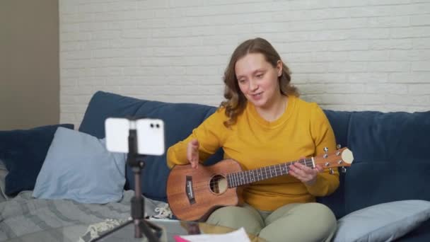 Mujer lleva a cabo lecciones en línea que enseñan a los estudiantes cómo jugar el ukelele. Se lo dice a la gente a través de un enlace directo de video. Ella explica cómo batir las cuerdas del instrumento. Blogging, capacitación en línea — Vídeos de Stock